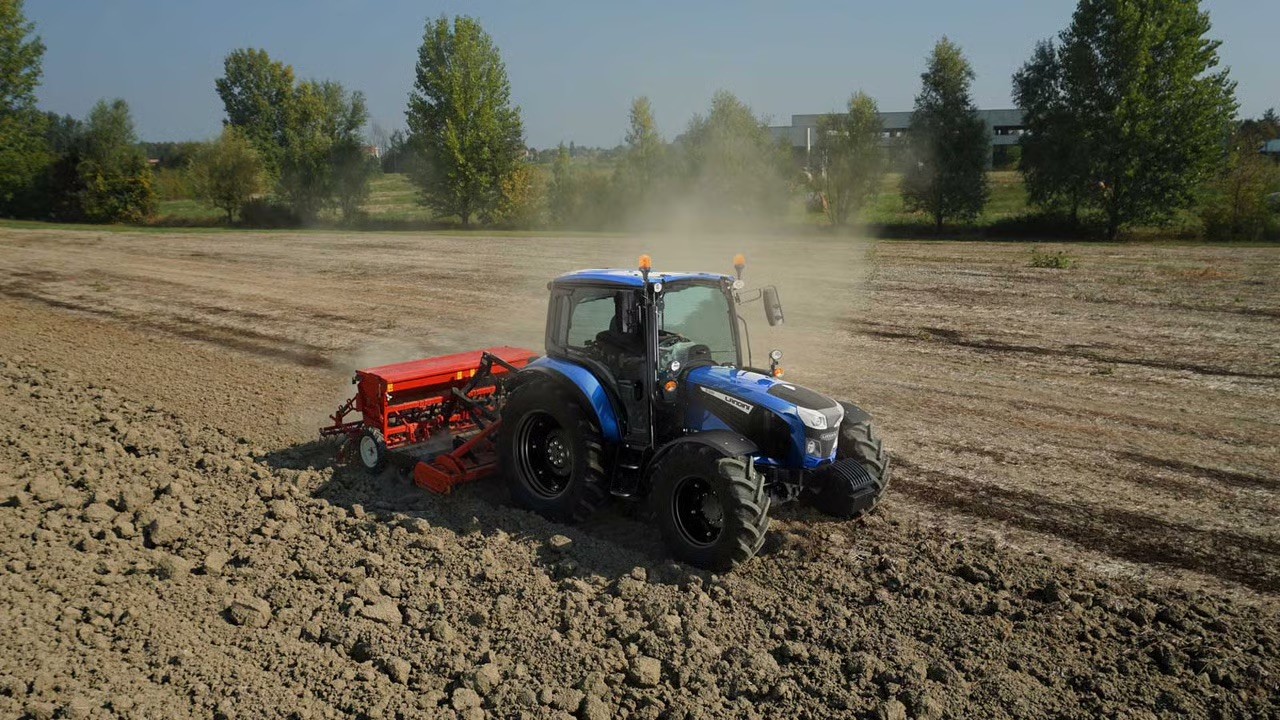 Blue tractor pulling a red seed drill across a dusty field
